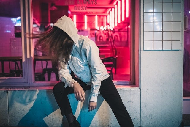 A woman in a hoodie, sitting a a business’s open window, leans away from view. A sign over her head says, “Always busy.”