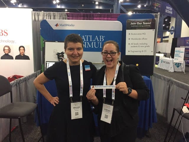 Two women in all black with white badges stand in front of a MathWorks booth at Grace Hopper Conference. There is a blue banner in the background that has the MathLab logo and there’s grey pipe and drap on each side.