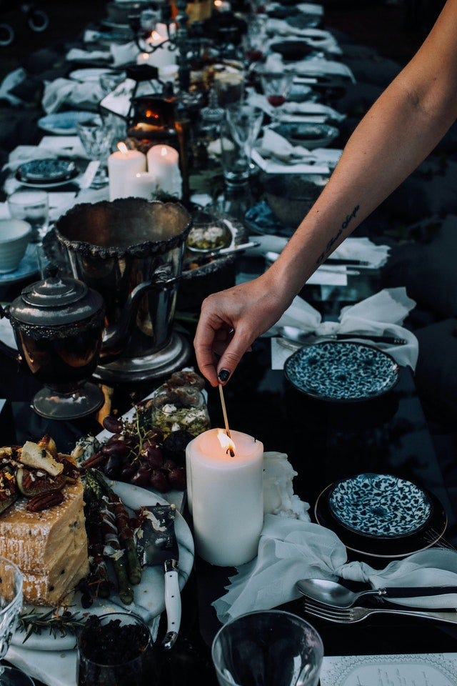 A table full of dinnerware and a feast of food fills the photo. In the foreground, an arm is extended through the center of the picture lighting the wick of a candle.