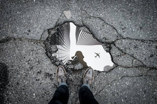 Photo Credit: Unsplash photo by Mark-Olivier Jodoin. This is a black and white photo of a buildings, airplance, and photographer in water on a cracked black top road.