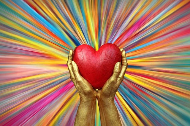 Gold sculpted hands hold a wooden red heart with rainbow colors beaming rays all around.