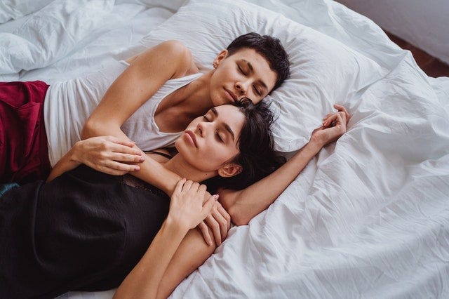 Two women sleep snuggled together in a bed of white sheets.