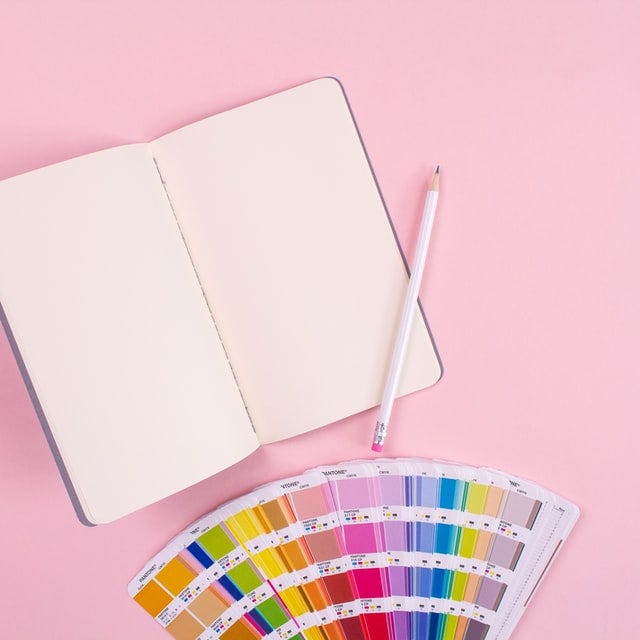 An open notebook and a pencil on a pastel pink background with a Pantone chip fan laid out below