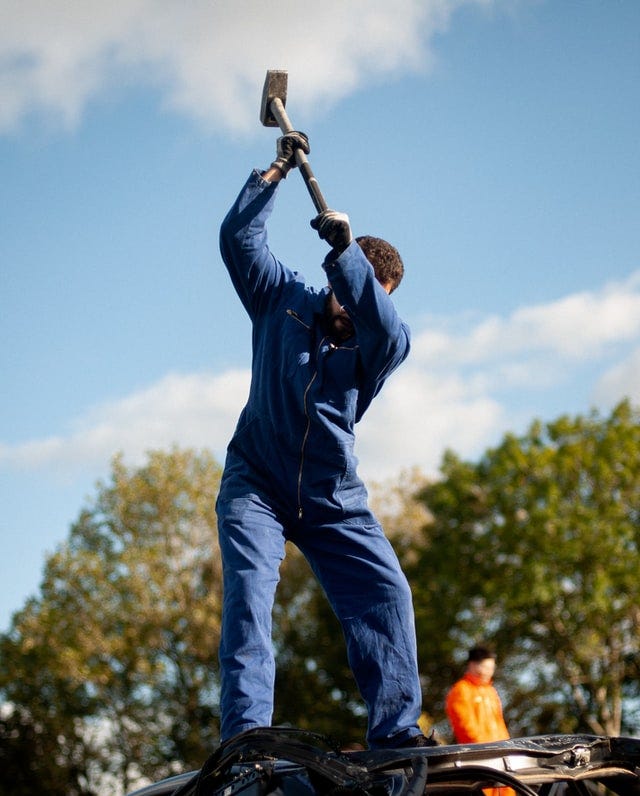 A person with a sledge hammer busting up the top of a car.