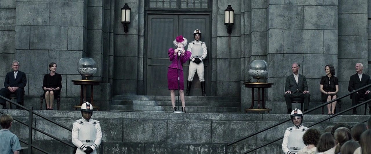 Effie Trinket’s vibrant wardrobe and make-up juxtaposes the drab clothing worn by the inhabitants of District 12.