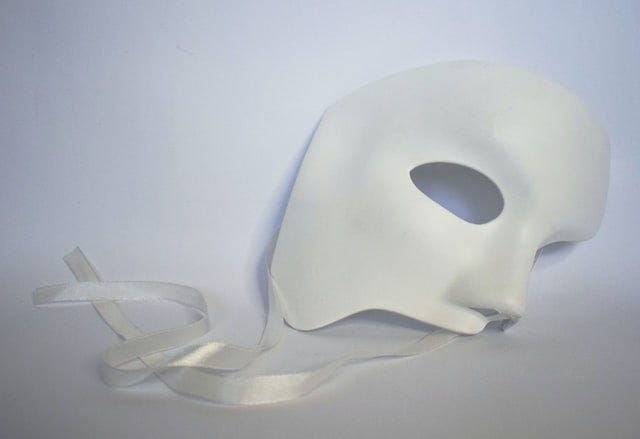 a phantom of the opera style mask lying on a white cloth with a swirling ribbon at the bottom.