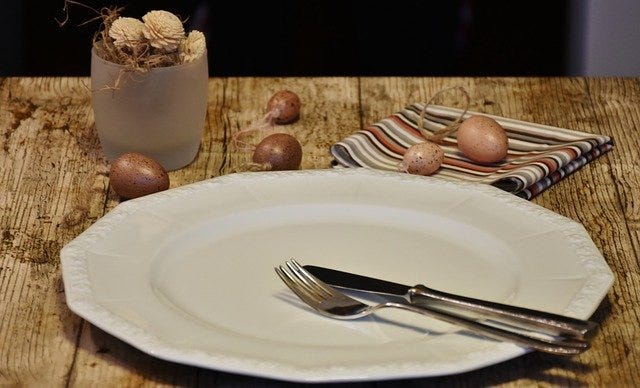 an empty plate, fork, knife, napkin, and vase sit on a wooden table