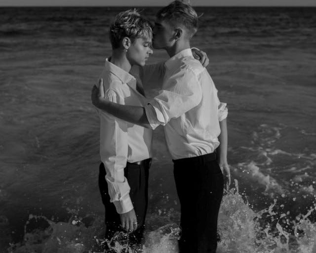 Two men standing in the water with their arms around each other. One man is kissing the other mans head.