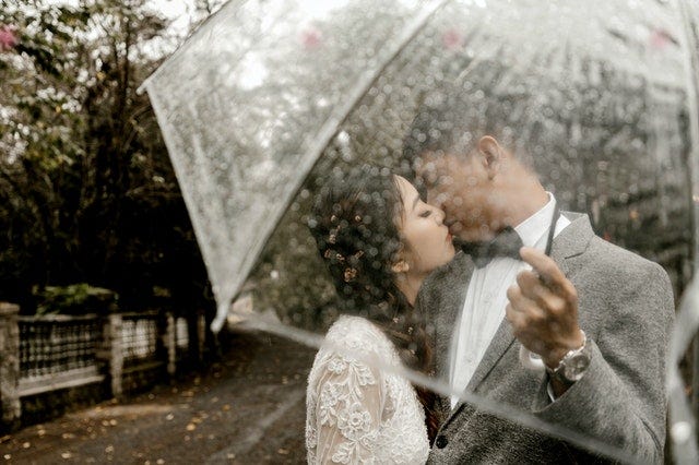A couple dressed in finery kisses beneath a translucent umbrella while it rains around them. Behind them is a walking path, trees, and fence.