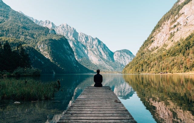 A person sits at the end of a long pier, looking out at the still water of a lake and mountains. The sky is blue and clear, it evokes a contemplative feeling.