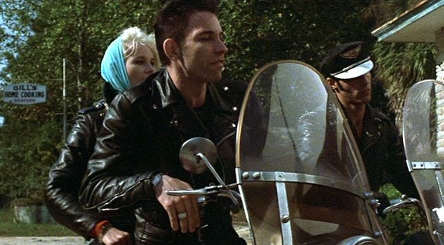 Robert Gordon rides a motorcycle with a blonde woman on the back, alongside a fellow biker gang member.