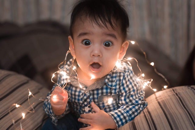A toddler wrapped in white holiday lights with a suprised look on his face.