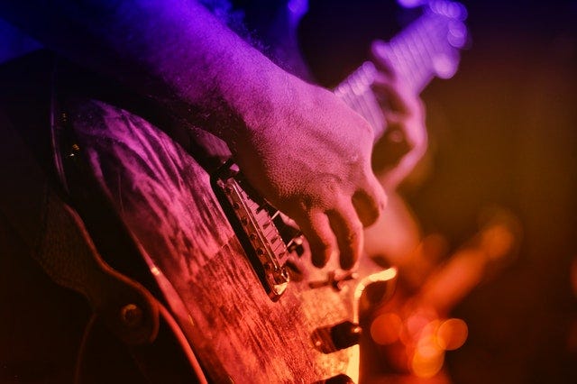 View of a hand strumming chords on an electric guitar as the other hand holds strings down. The guitar and hands are bathed in a rainbow of stage lights.