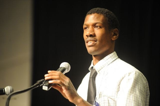 Max Barrows, a Black man wearing a white checked shirt and a black tie with his hand on a microphone, presenting at the SABE conference