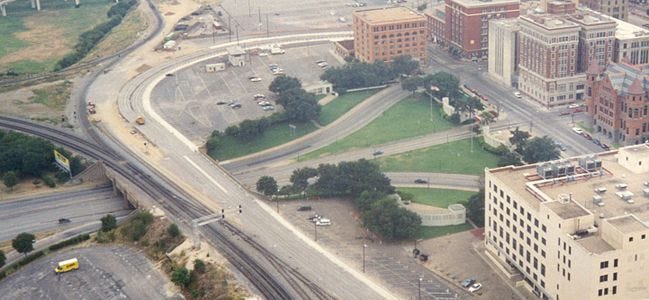 Aerial shot of Dealey Plaza in Texas, showing a number of intersecting roads as traffic approaches large square buildings.