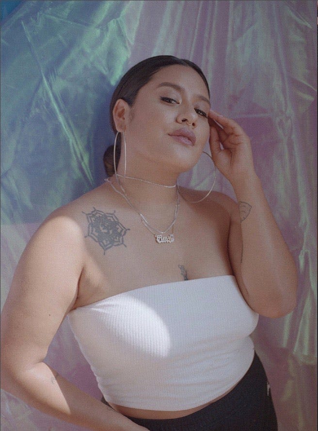 Amy Quichiz posing with her fingers held to her left temple in front of a backdrop of iridescent fabric.