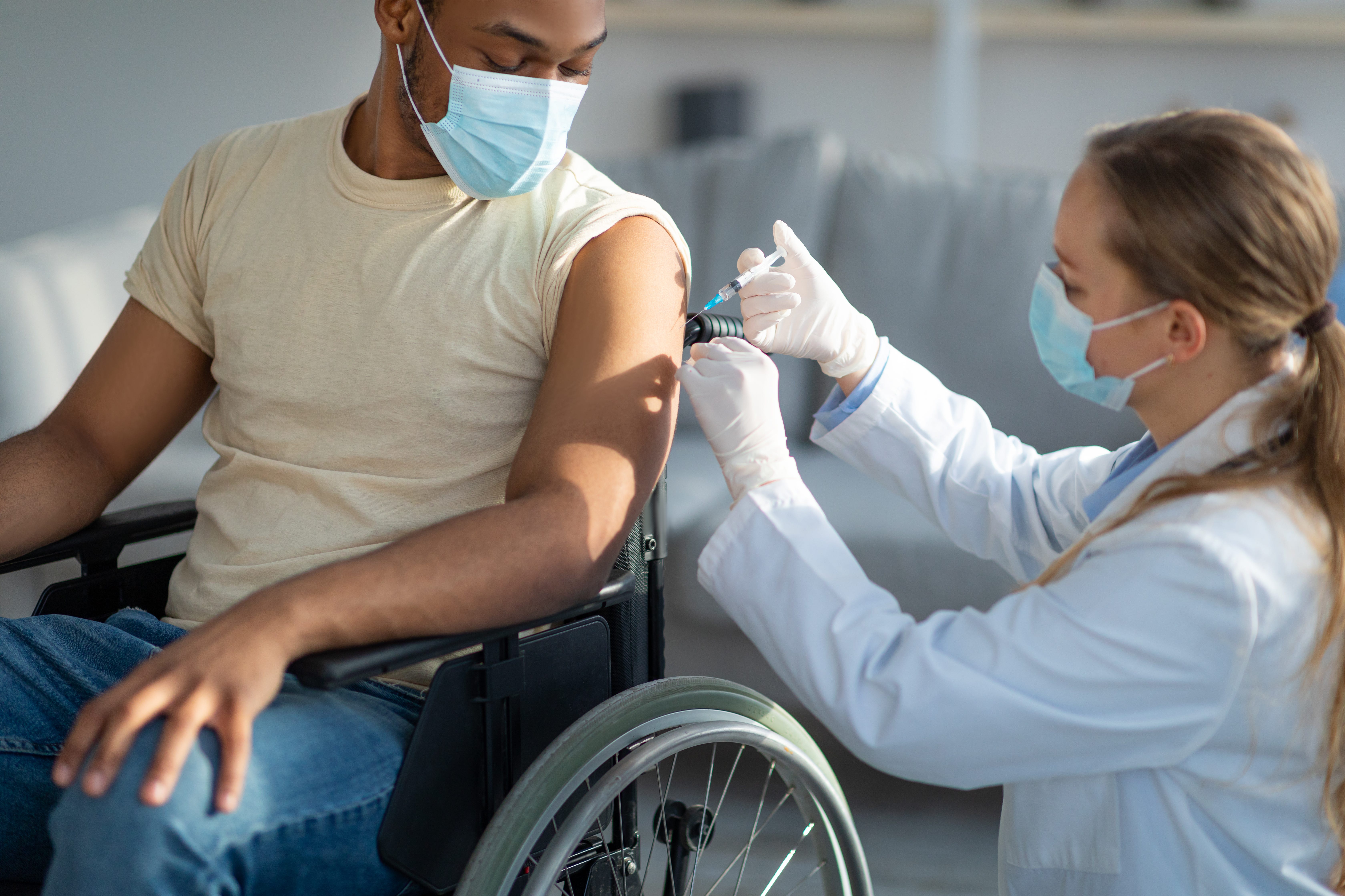 Kneeled doctor giving coronavirus vaccine to a masked patient sitting in a wheelchair.
