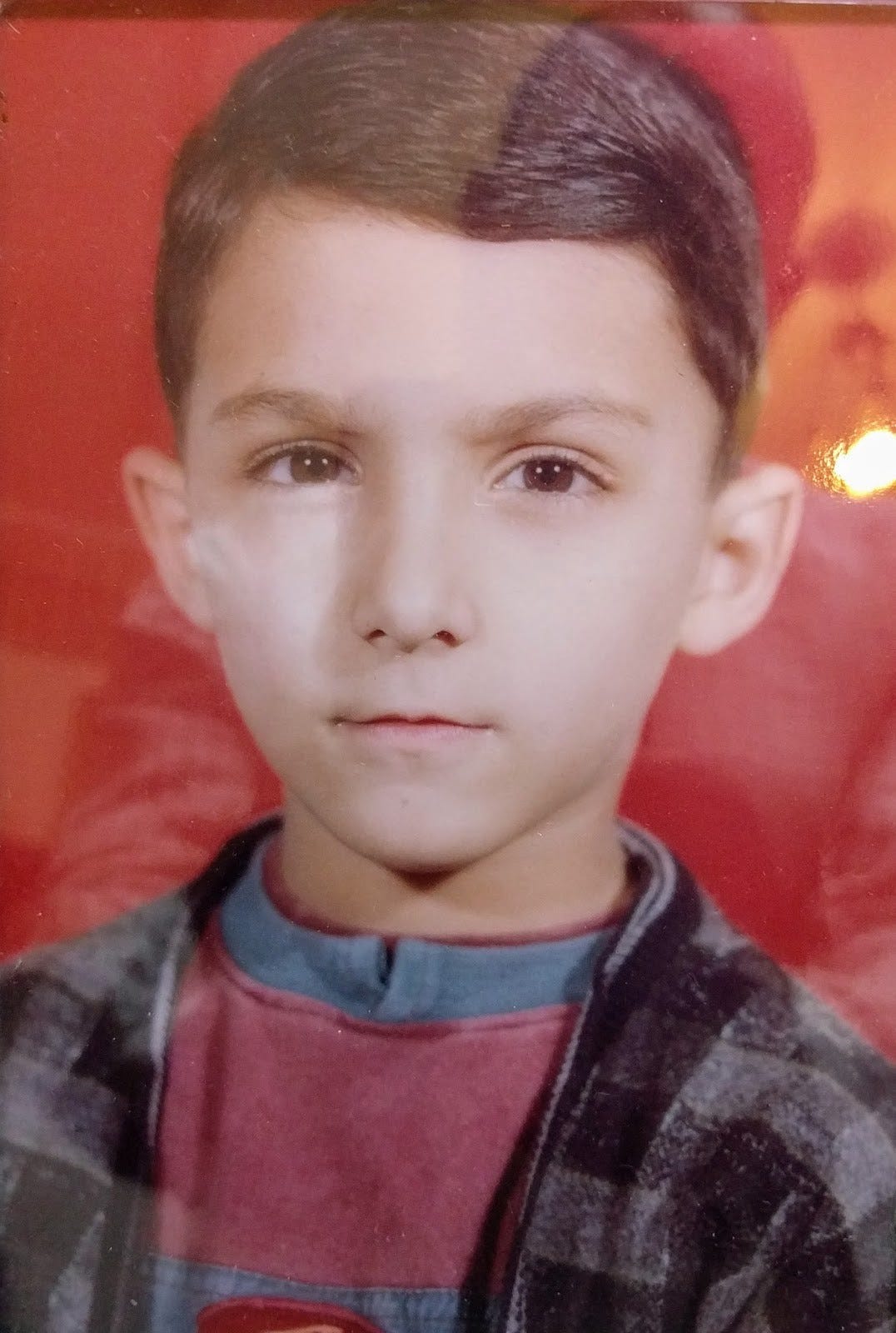 Portrait of child me with a serious expression, wearing a red top and a checkered grey cardigan, against a red background