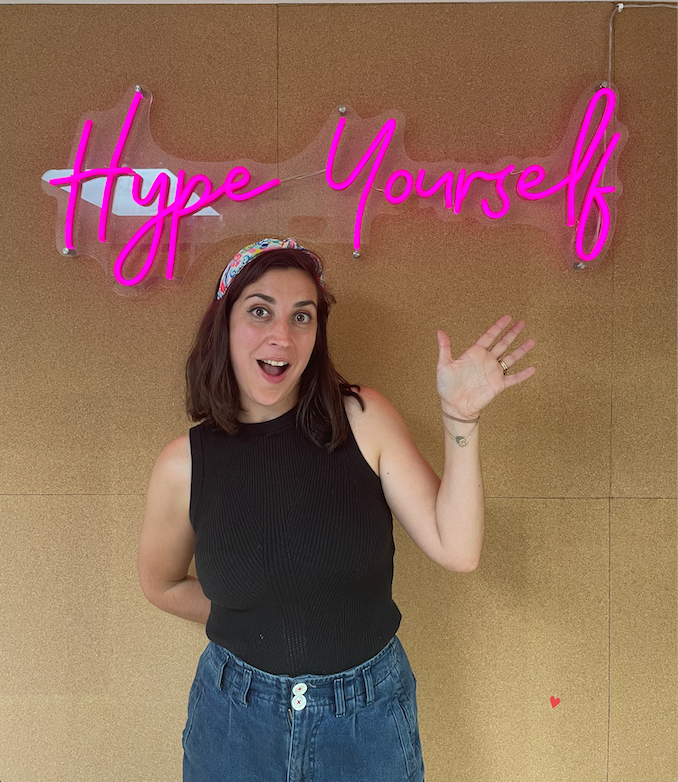 Picture of author Lucy, waving in front of a pink neon sign that reads Hype Yourself.