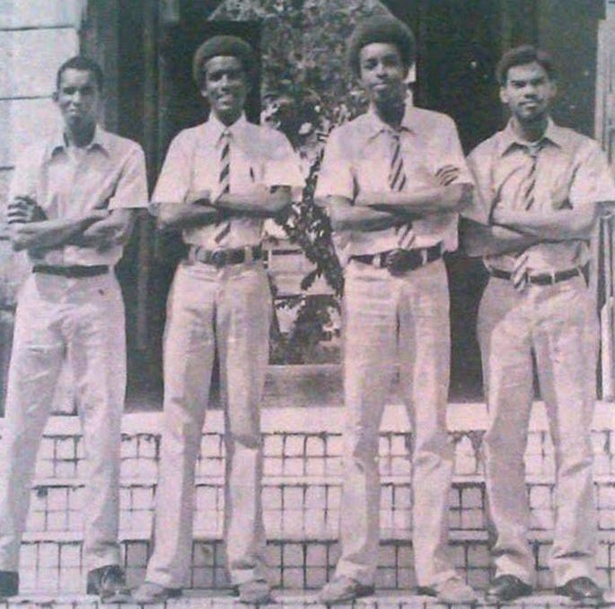 Four Black teenage boys standing with arms folded, each wearing a shirt, long pants and tie.