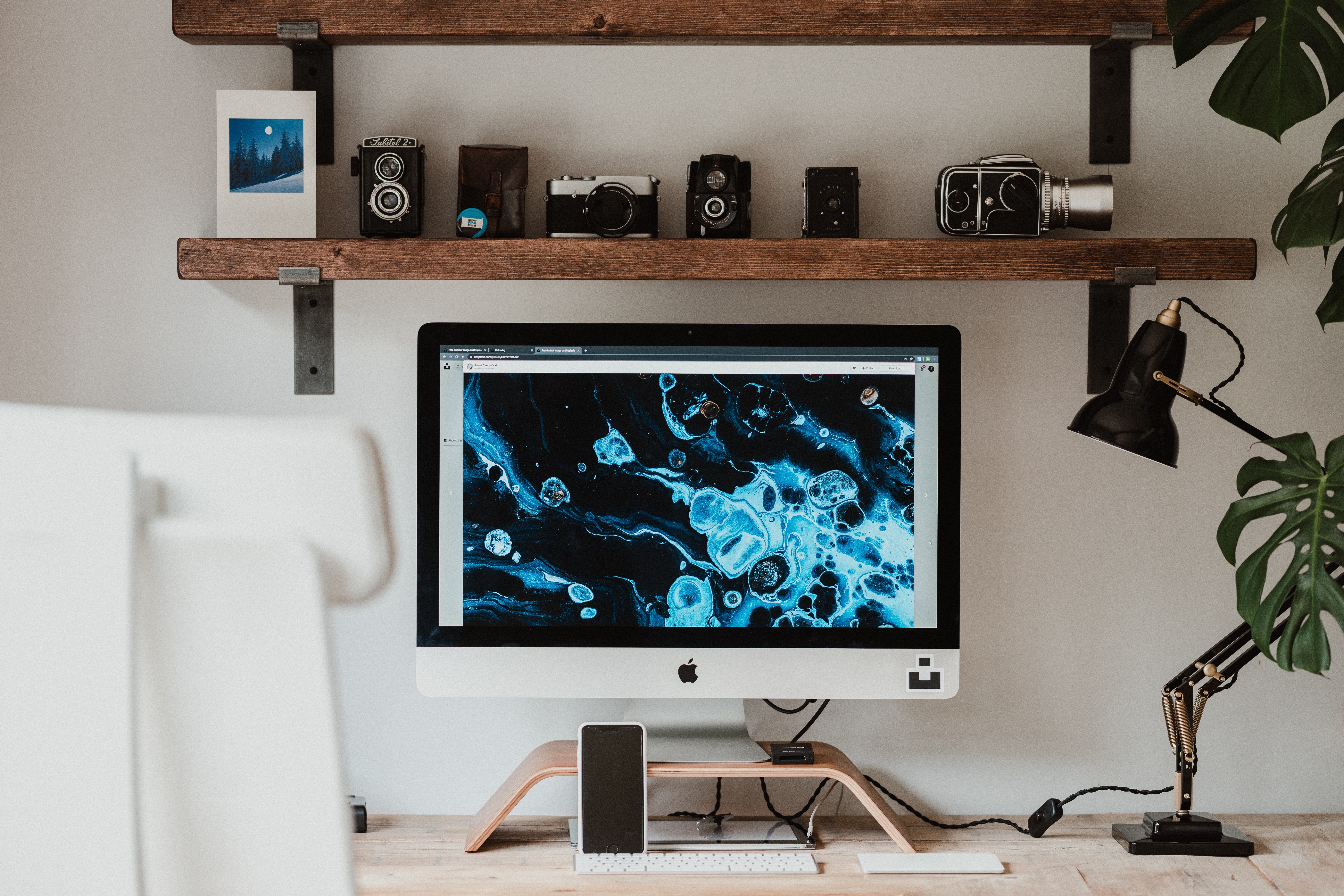 An Apple desktop on a table with a lamp, shelves, and a few Monstera leaves.