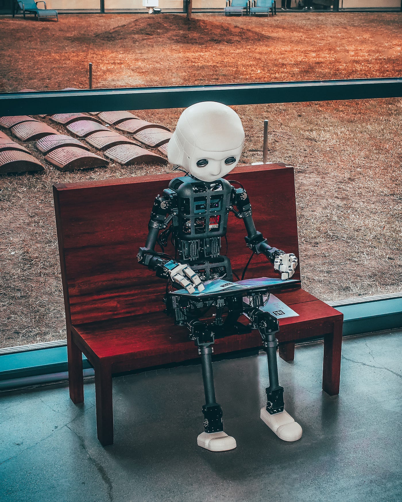 A robot sits on a small wooden bench, typing at a keyboard. The robot’s head is white and solid, while we see his uncovered body’s circuitry.