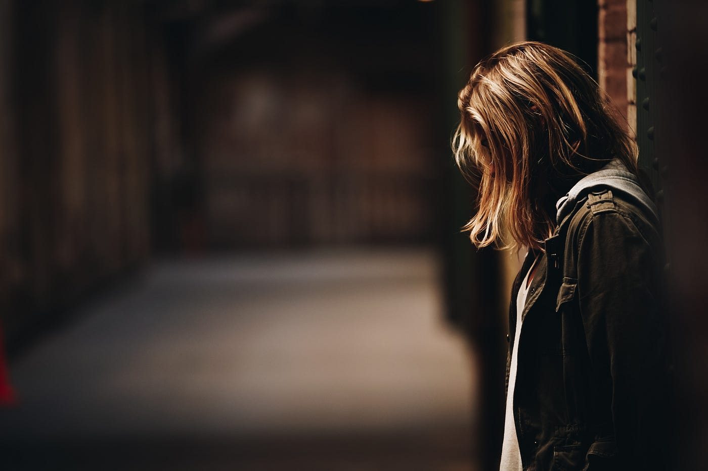 Young woman, with sandy brown hair, stands in profile to the right of the image. She faces left and appears to be in a poorly lit alleyway.