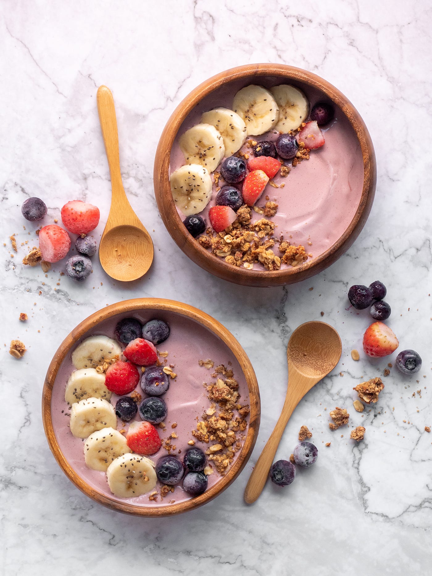 Two bowls of yogurt, with banana slices, raspberries, and blueberries. The yogurt is pink and we see it all from above. There are two wooden spoons, one next to each bowl.