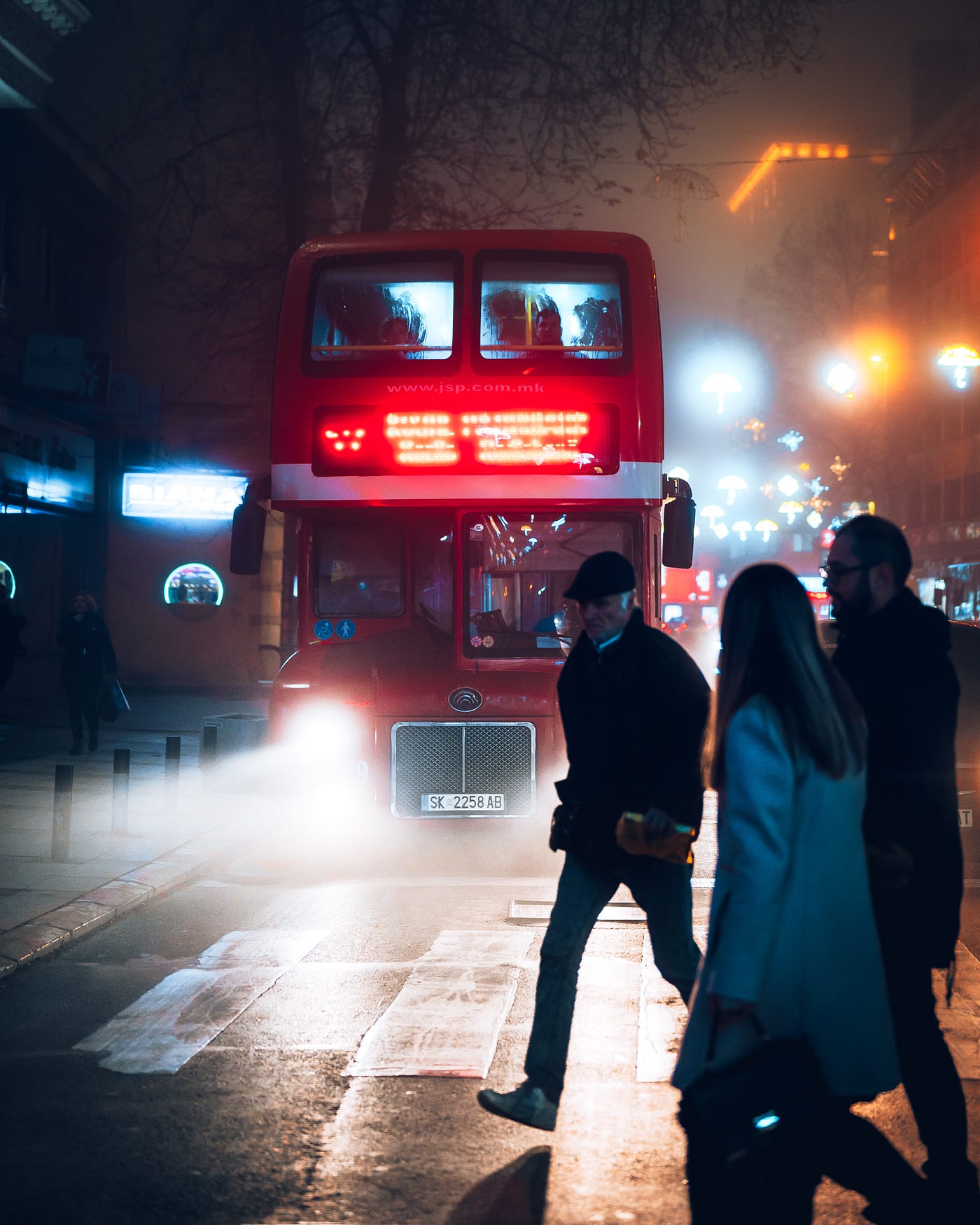 Three individuals walk in front of a London double decker red bus at night.