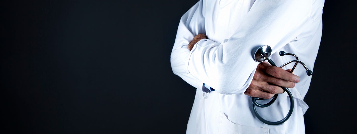 A soctor in a traditional white jacket, arms crossed and a curled stethoscope in his/her right hand. We see the clinician from waist to upper chest level. There is a black background.