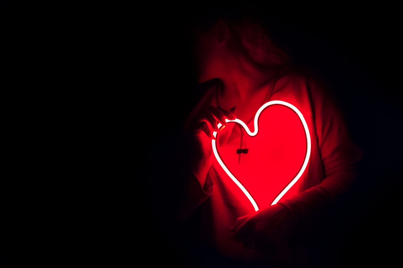 A woman holds a heart-shaped neon wire against her chest. Dark scene, with only light from the neon.
