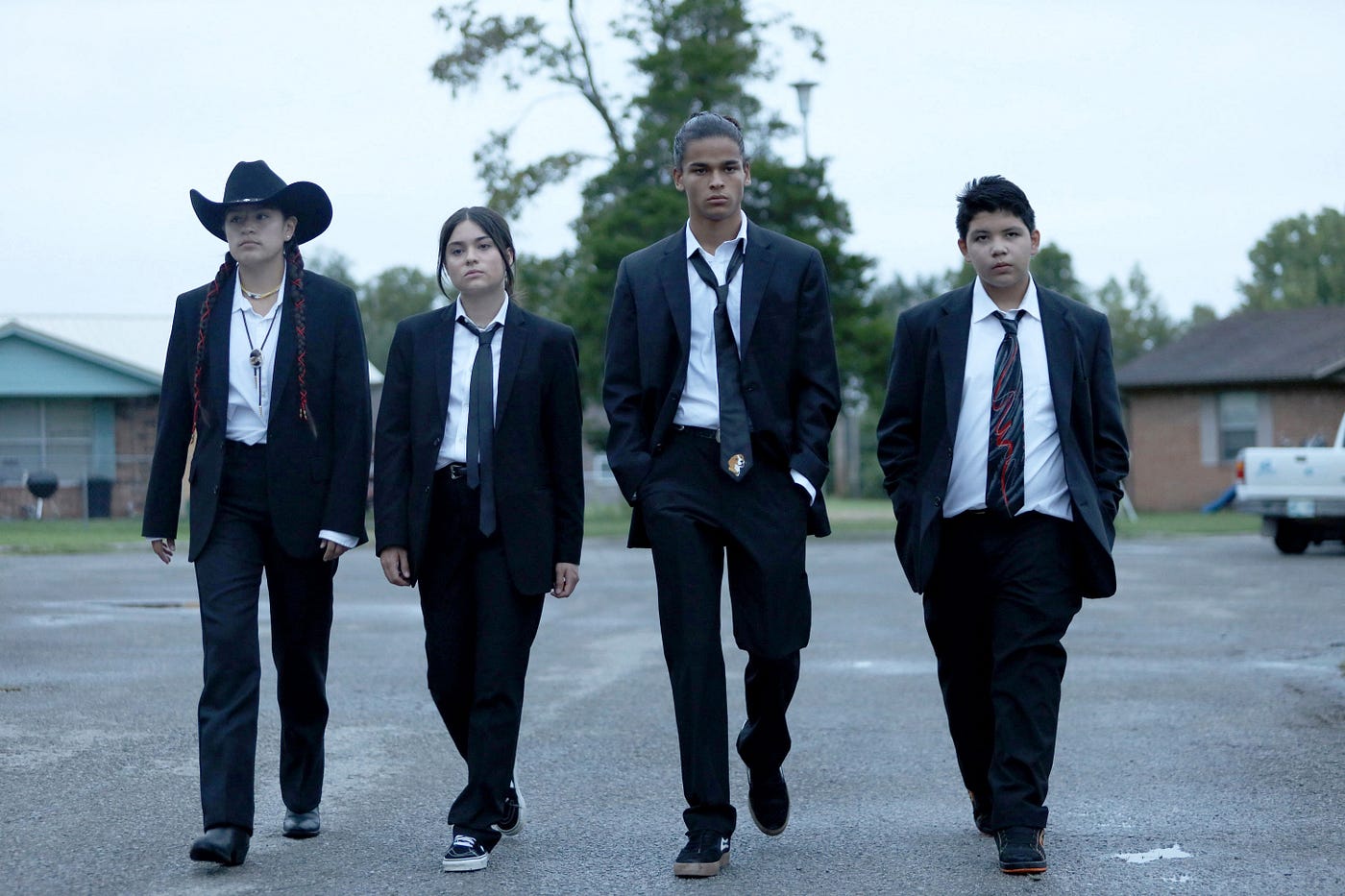 The titular reservation dogs, from left to right Paulina Alexis as Willie Jack, Kawennáhere Devery Jacobs as Elora, D’Pharaoh Woon-A-Tai as Bear, and Lane Factor as Cheese, walk along the street towards the camera in black suits in the manner of Reservoir Dogs from the 1992 Quentin Tarantino film.