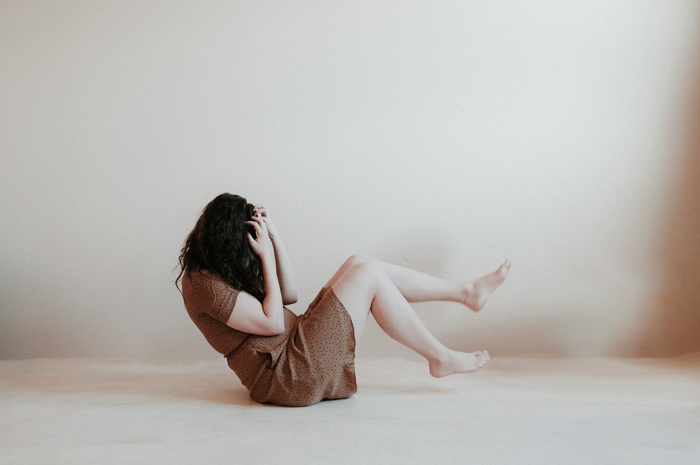 Woman sits on the floor, her head (with hands over her face) to the left, and her legs (lifted off the floor) to the right. She wears a brown simple cotton casual dress. Off-white background, off-white floor.