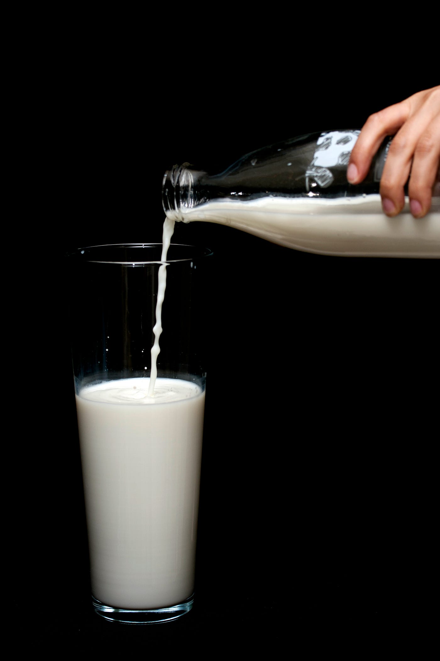 Someone extends a hand from the right to pour milk into a glass from a glass bottle.
