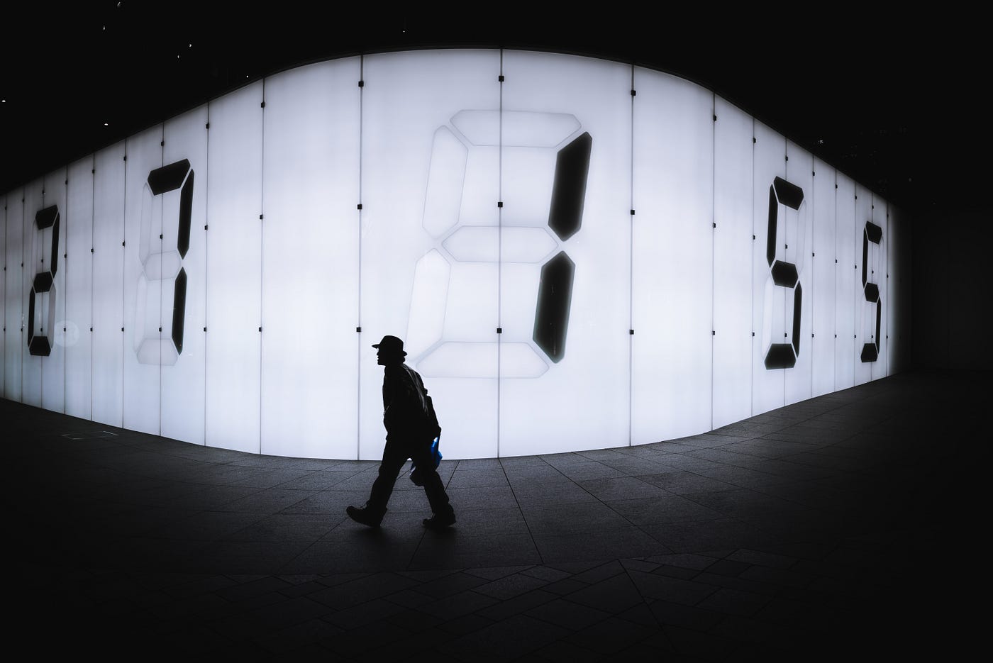 A black and white image of a man (in silhouette) walking from right to left. The man is in the middle of the image, and behind him is a wall of square panels with numbers on some of them.