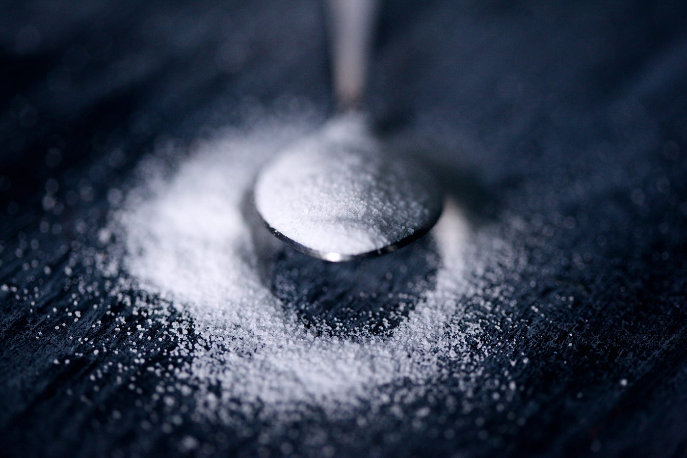 A spoon extends from the image top, a bit of sugar in it, but most of the sugar has spilled out onto a dark gray surface. Table sugar is composed of glucose and fructose.