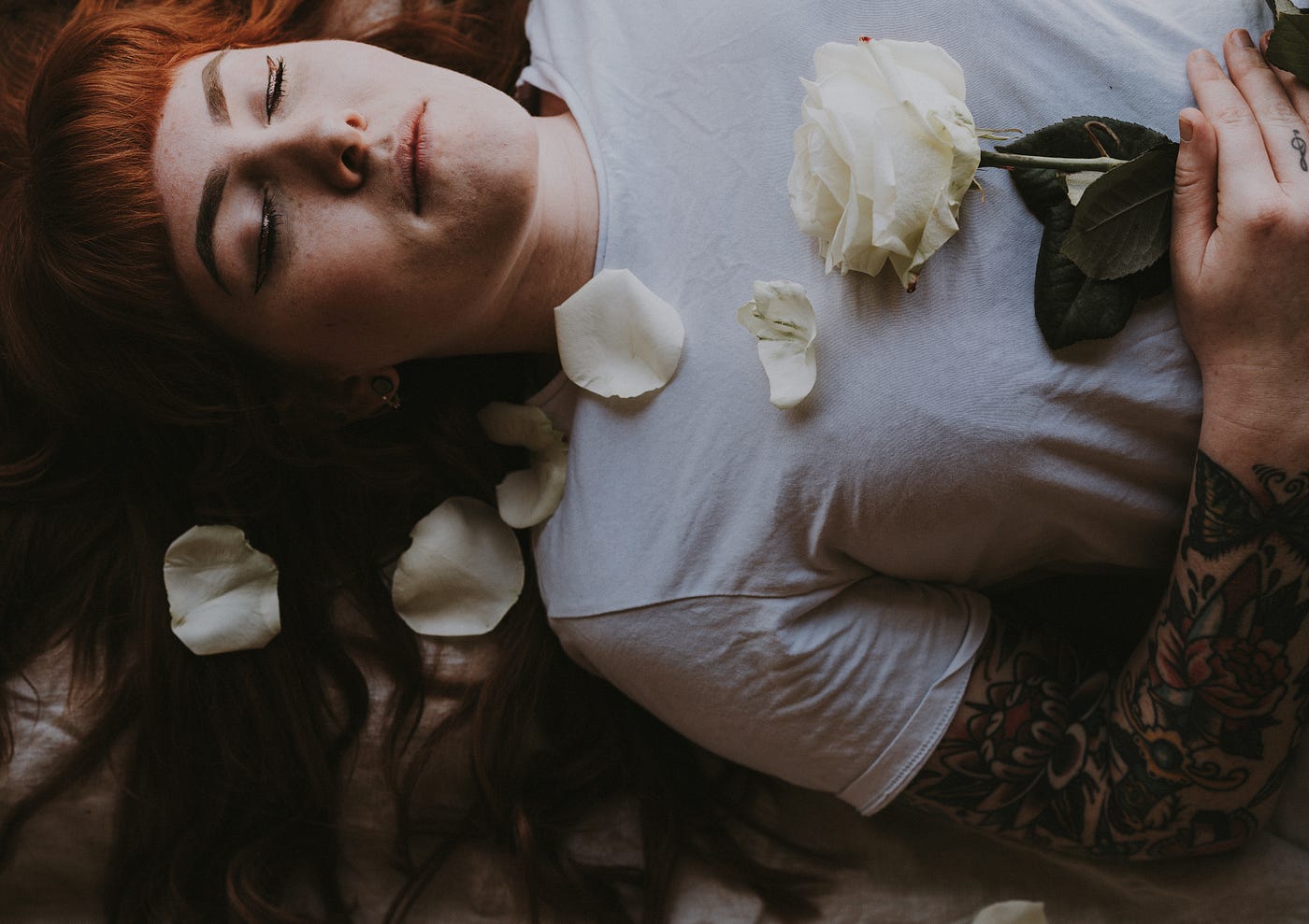A red headed, fair skinned young woman lays eyes-closed on the ground, her hands holding a white rose, whose petals are scattered across her chest.