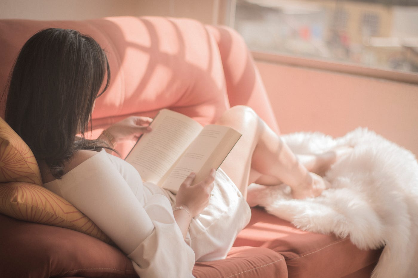 woman reading a book on a pink couch with sunlight gently streaming in.