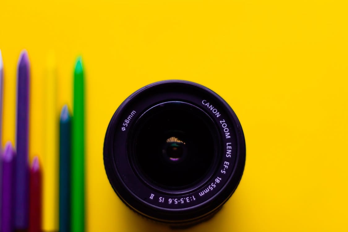 A picture of a camera lens on a yellow table with blurred out pencils beside it.