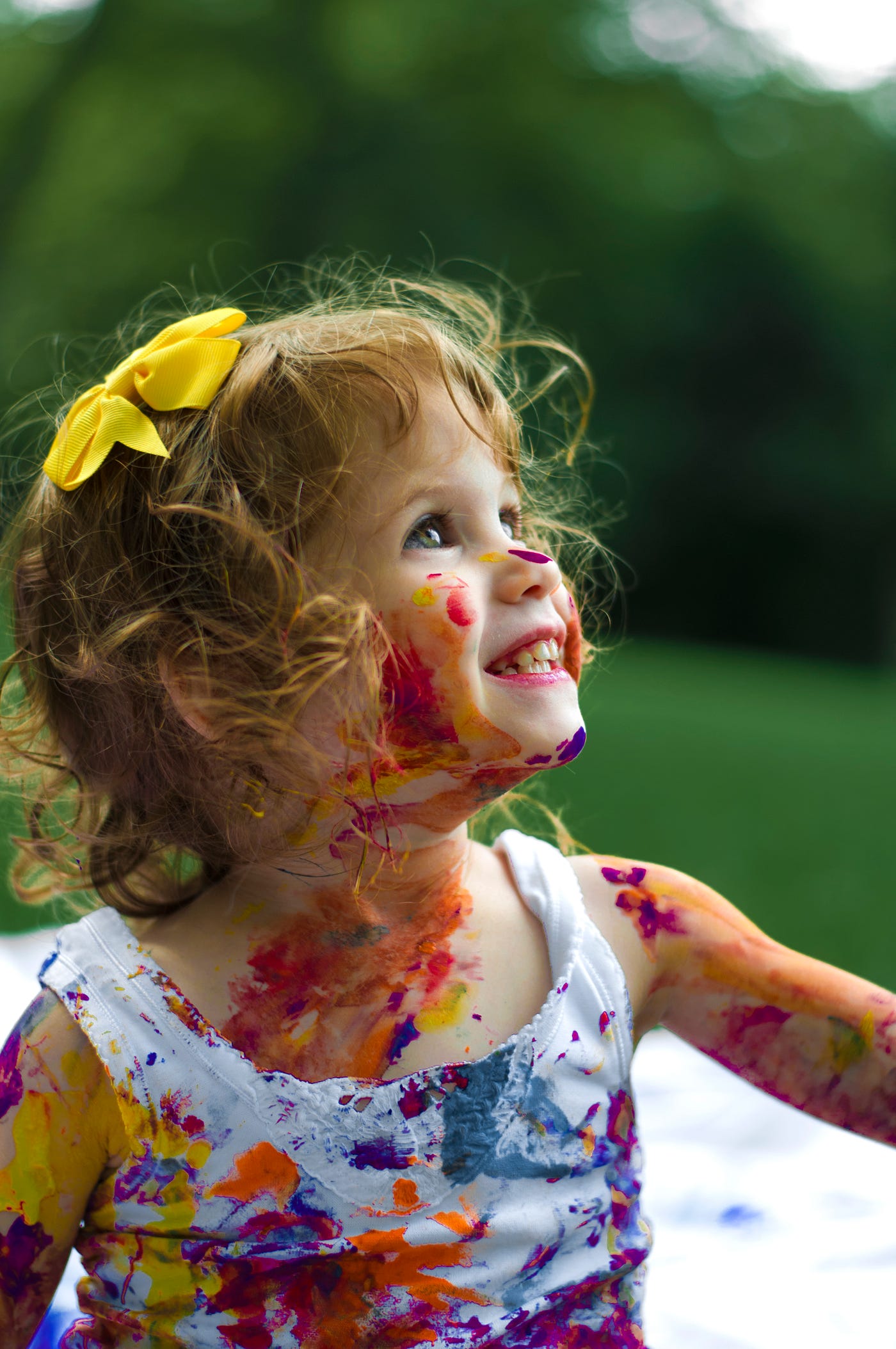 White child, around age 2 or 3, smiles as she gazes upward. She has curly reddish hair, a yellow ribbon in her hair, and various colors of pain splashed onto her lower face and neck. We see her in profile as she faces to the right.