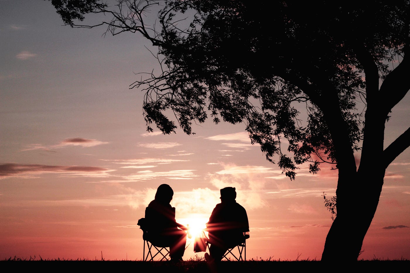 An elderly couple sits in lawn chairs under a tree, facing away from the camera. The sun sets in the background.