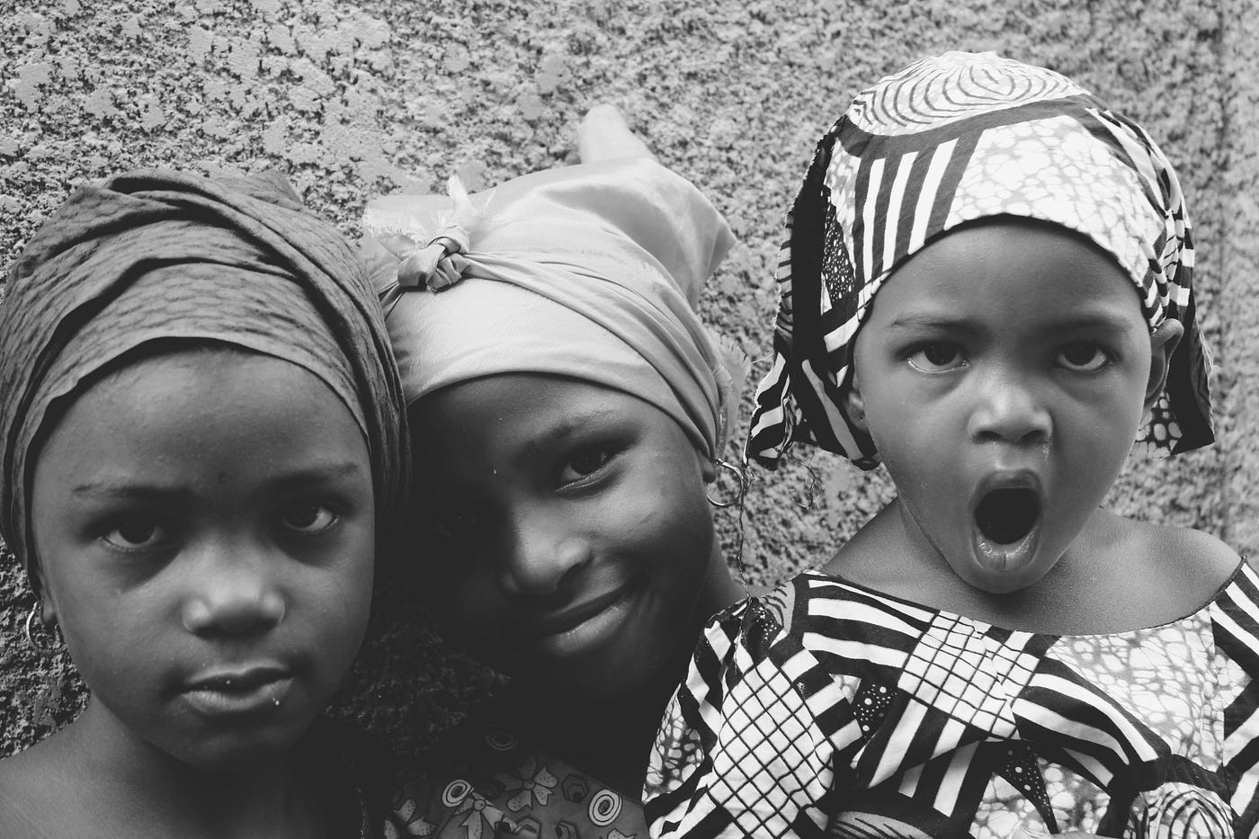 Three young children of color, wearing traditional scarves, look at the camera.