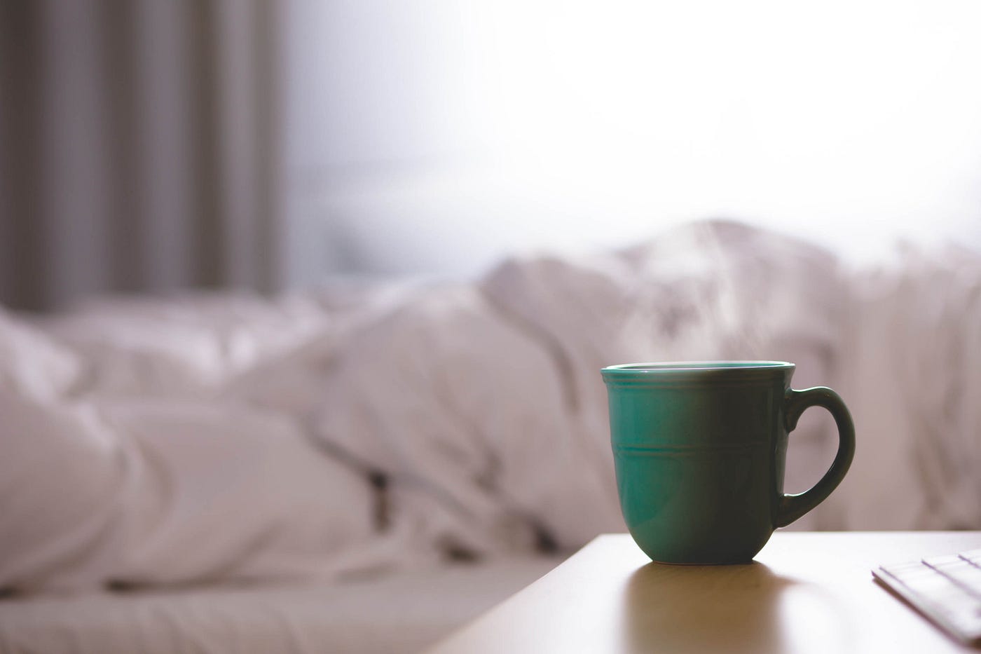 A green coffee cup (with its oval handle to the right) sits on a wooden desk in the right foreground. Steam rises from the cup. A messy bed appears blurred in the background.