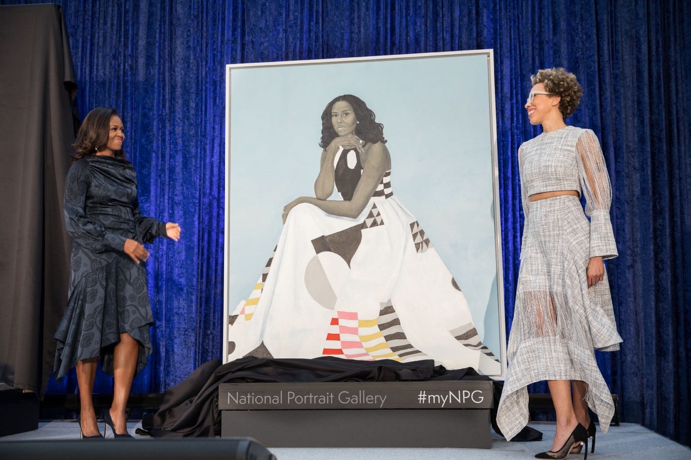 Michelle Obama and Amy Sherald stand on stage on either side of a large portrait of Michelle.