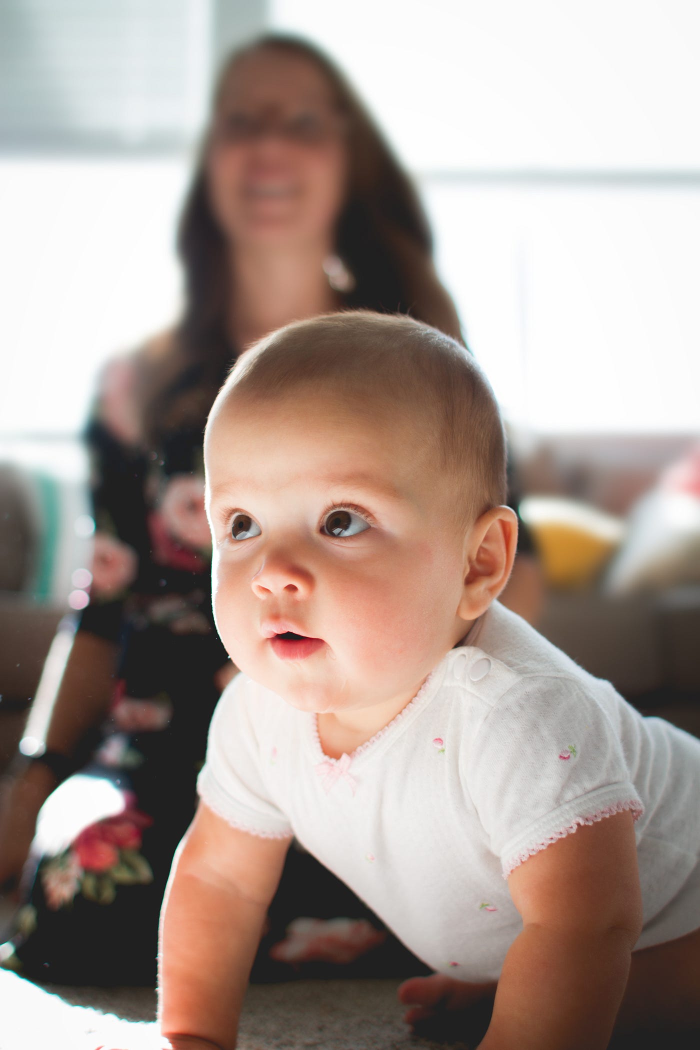 A cute white baby perches up on his arms to look towards the front/right. A woman sits in the background.