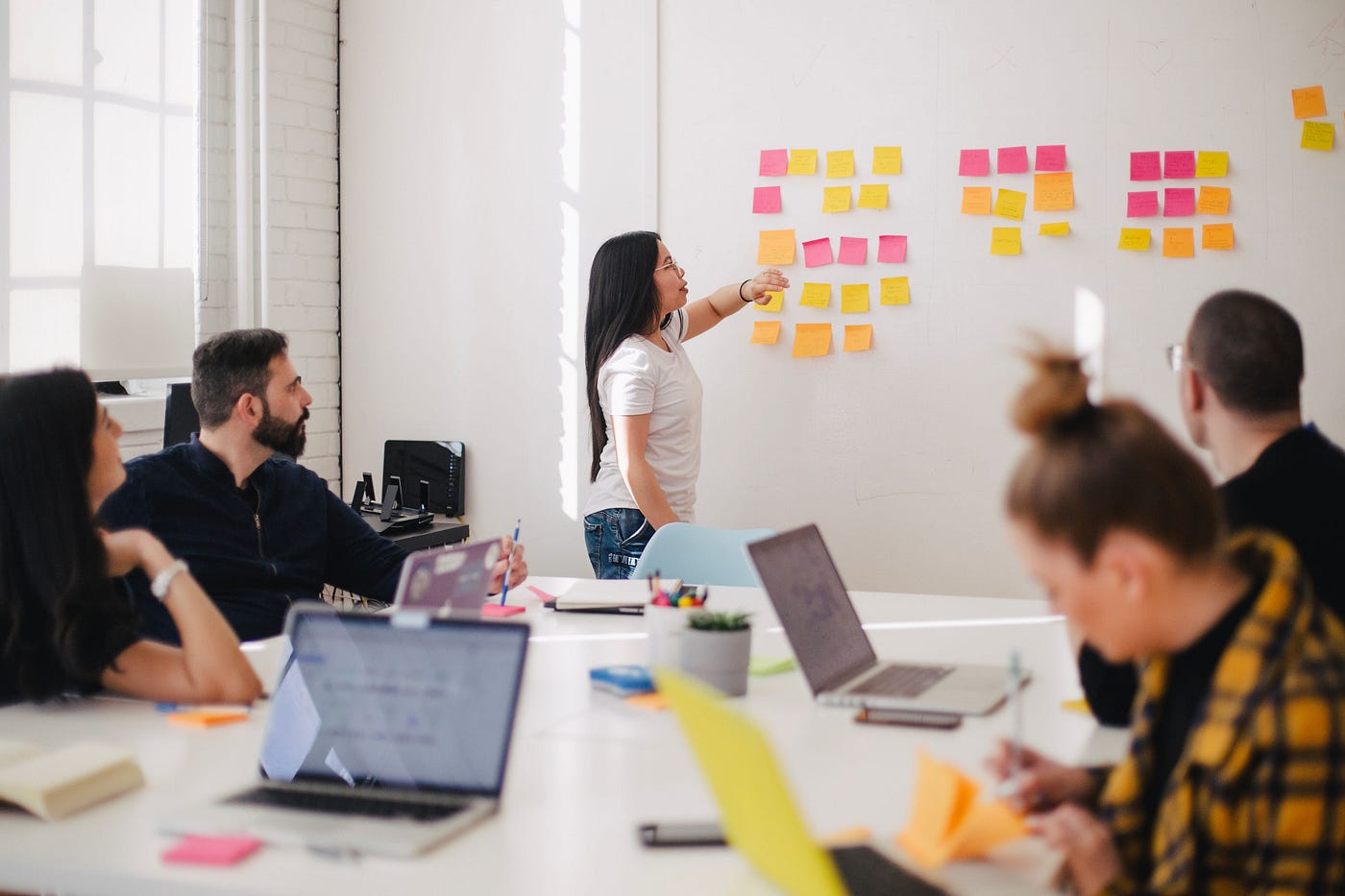 A woman standing in front of a white wall with post-its on them, while a group of young professionals with laptops watch her.