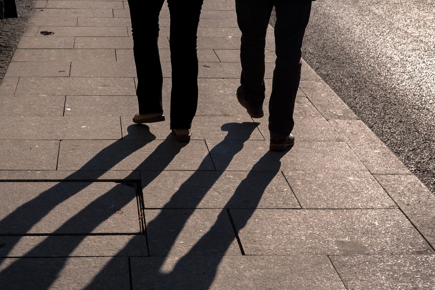 We see two sets of legs walking away from us on a sidewalk with pavers. Nothing visible from upper legs up. Shadows come off each of the legs, towards us. Black and white image.
