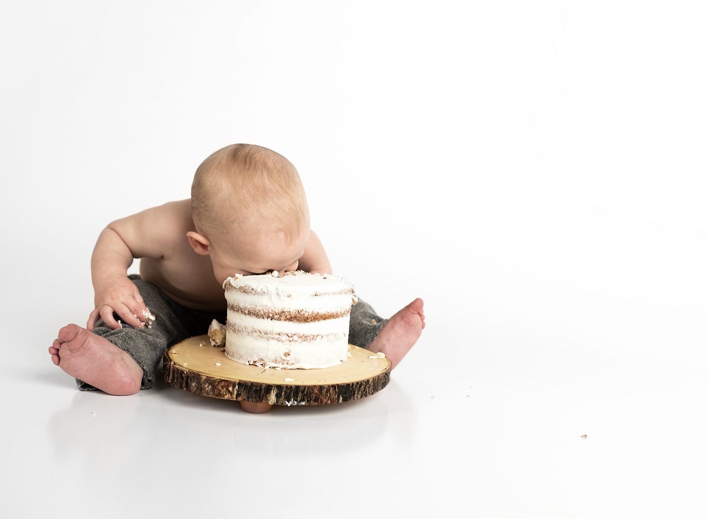 A toddler puts his face into a white cake, the dessert between his splayed legs (as he sits).