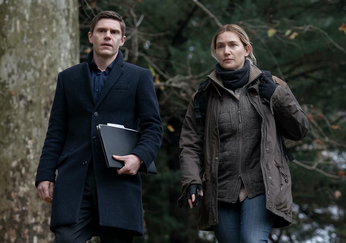Two characters dressed for cold weather walk towards the camera in the woods. From left to right they are Colin Zabel played by Evan Peters and Mare Sheehan played by Kate Winslet.