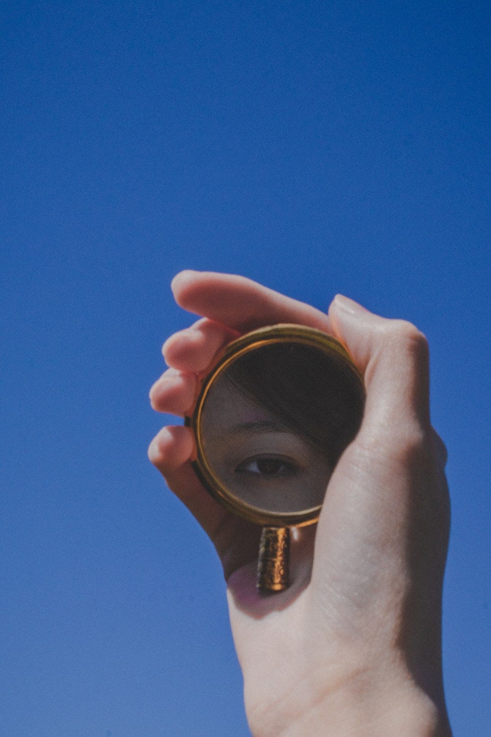 Woman holds up small mirror against blue sky. Her brown hair and a brown eye is shown in the reflection.